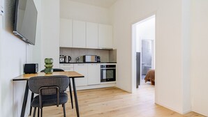 Cocina compacta con armarios blancos, microondas y estufa. Hay un comedor con mesa y sillas, y un televisor montado en la pared.