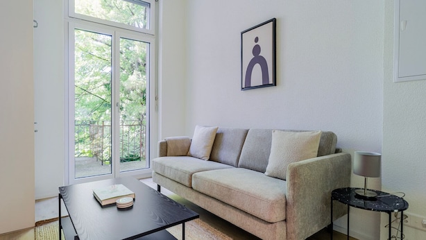 Una sala de estar moderna con un sofá, una mesa de centro y una lámpara. Hay un ventanal con vistas a zonas verdes.