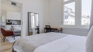 Een slaapkamer met een bed, een bureau, een stoel, een spiegel en uitzicht op gebouwen.