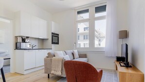 Een compacte leefruimte met een kitchenette, een bank, een rode stoel, een televisie en een raam met uitzicht op gebouwen.