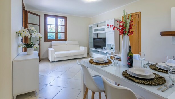 A modern living room with a white sofa, a dining area with a glass table, and a kitchen with white cabinets.
