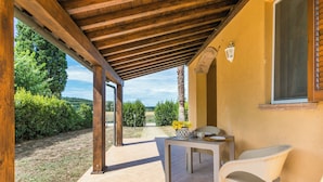 An outdoor patio with a wooden pergola, a table with chairs, and a window overlooking a garden.