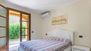 A bedroom with a large bed, a small bedside table, a sliding glass door leading to a balcony, and a wall-mounted air conditioner.