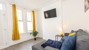 A living room with a grey sofa, yellow curtains, a flat-screen TV, and a small table with a lamp.