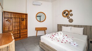 Habitación de hotel con un armario de madera, una mesita de noche pequeña de madera, un espejo redondo y una cama con ropa blanca y pétalos de rosa.