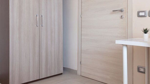 Una habitación con armario, una puerta, una mesita con una planta y un termostato fijado a la pared.