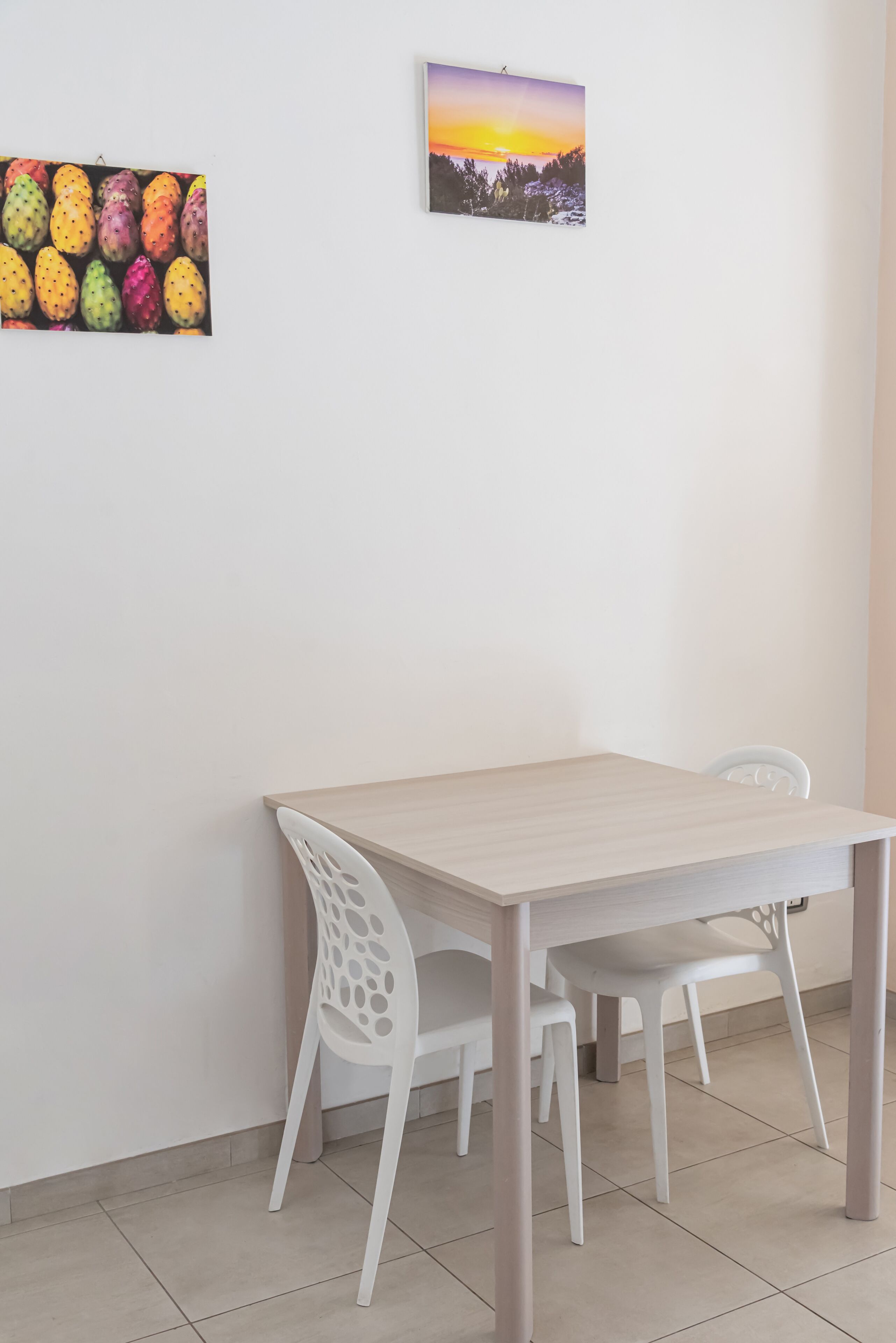 A dining area with a light wooden table and white chairs, two wall-mounted artworks, and a tiled floor.