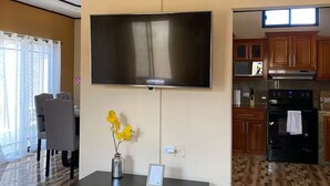 Un salon moderne avec un téléviseur à écran plat fixé au mur, une table basse noire ornée d’un vase de fleurs jaunes et un coin repas avec une chaise et une table grises.