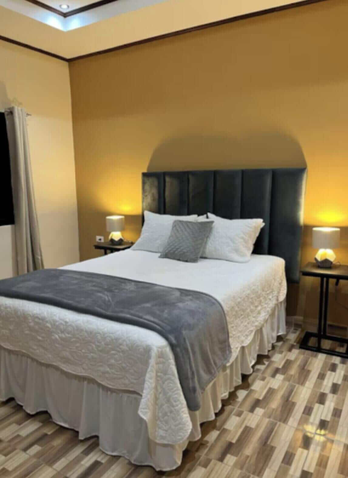 Un dormitorio ordenado con una cama grande, dos mesitas de noche con lámparas, una ventana con cortinas y suelo de madera.