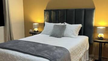 Un dormitorio ordenado con una cama grande, dos mesitas de noche con lámparas, una ventana con cortinas y suelo de madera.