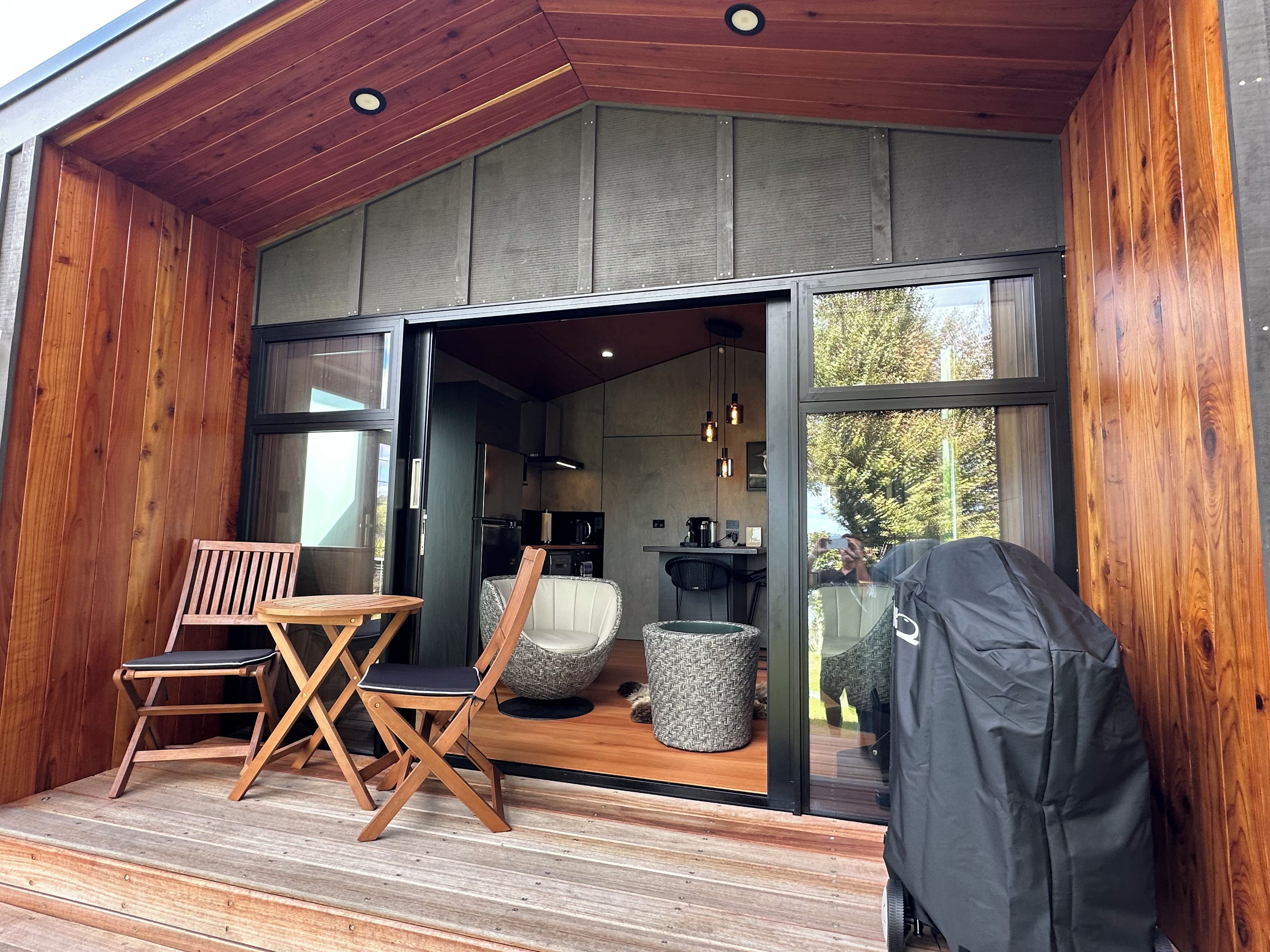A wooden deck with a dining table and chairs, a grill, and a view into a modern kitchen.
