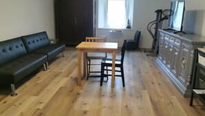 Un salon doté d’un plancher en bois, d’un canapé noir, d’une table à manger en bois avec des chaises, d’un téléviseur posé sur un meuble et d’une fenêtre laissant entrer la lumière naturelle.