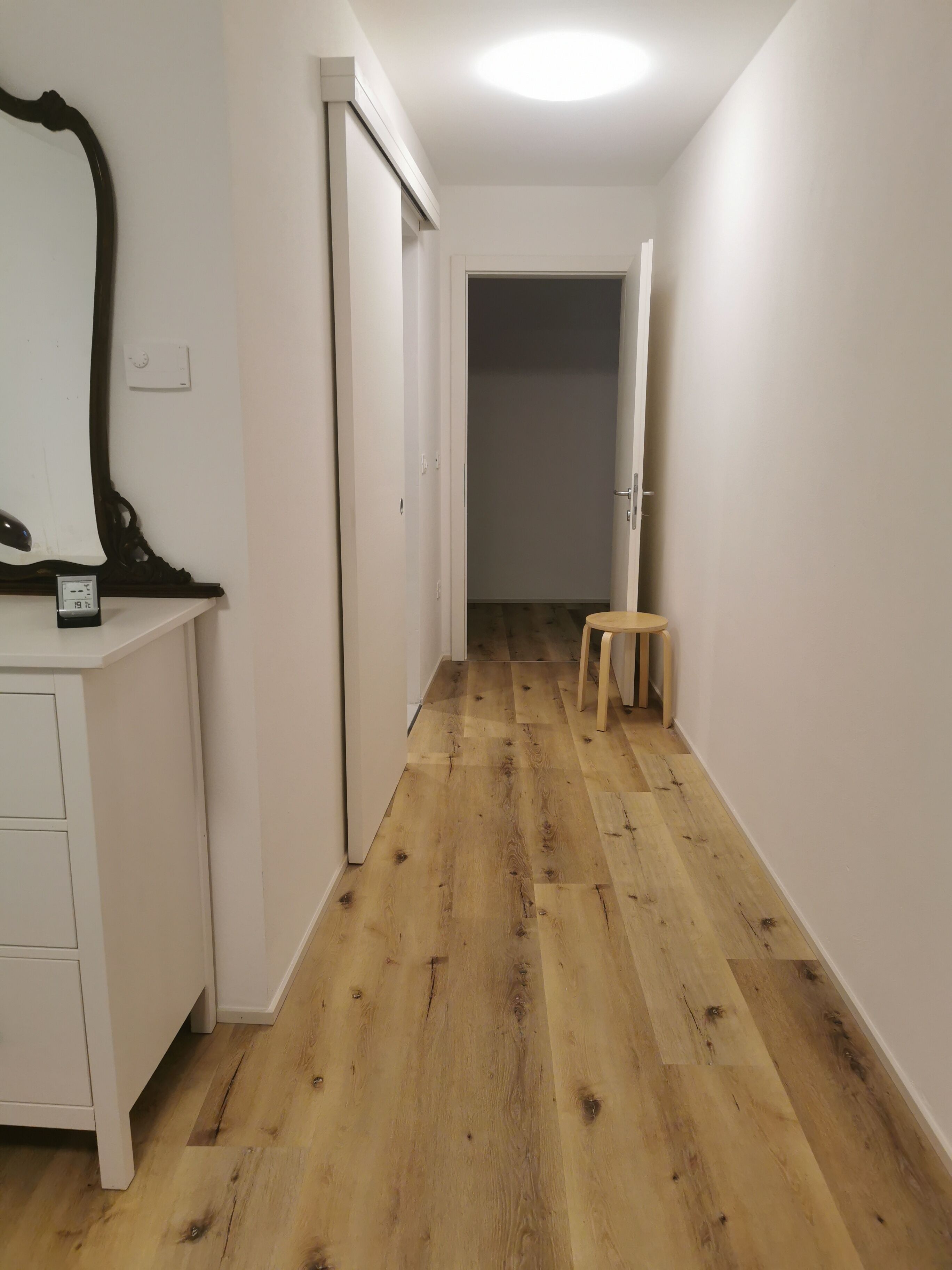 Ein schmaler Flur mit Holzboden, weißen Wänden und einem kleinen Hocker.