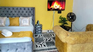 Una stanza con un letto, una televisione che mostra un fuoco, una ventola e una pianta.
