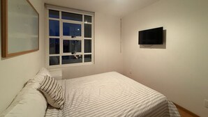 Ein Schlafzimmer mit Bett, Fenster, einem an der Wand montierten Fernseher und einem gerahmten Bild.