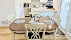 Uma sala de jantar com mesa de madeira e cadeiras brancas, uma cozinha ao fundo e uma janela com persianas.