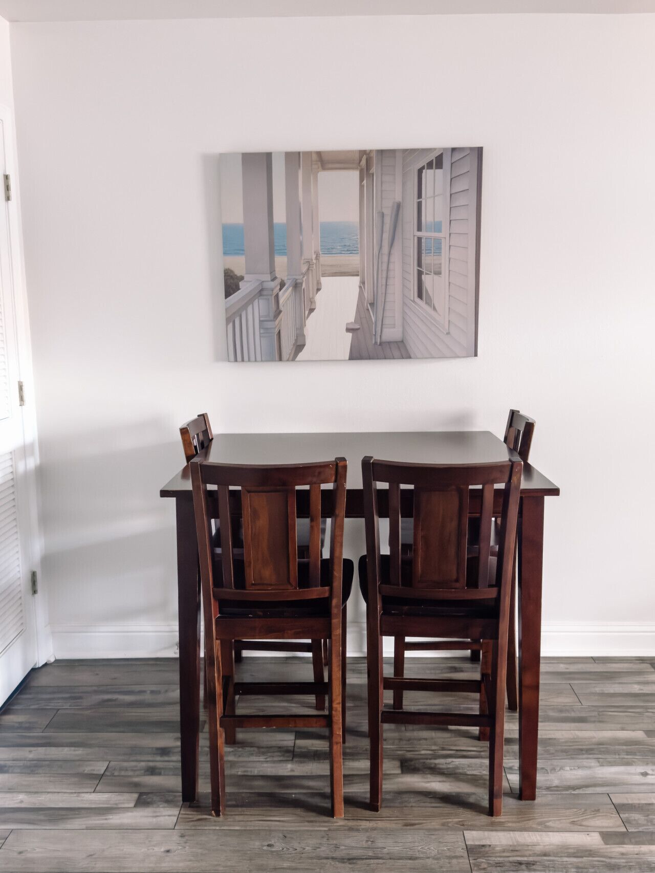 Un coin repas avec une table et des chaises en bois, un tableau représentant un paysage côtier et un plancher en bois.