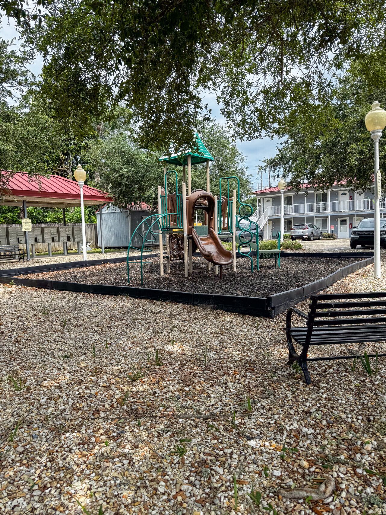Un parque infantil con un tobogán, rodeado de grava, bancos y árboles.