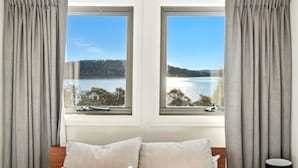 A bedroom with two windows overlooking a lake, a bed with pillows, and a bedside table.