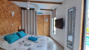 A bedroom with a wooden wall, a bed with floral bedding, two pillows, a nightstand with a lamp, a ceiling fan, a TV, and a window with curtains. - Private Bungalow ZANANAS (97435)