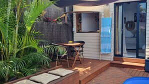A patio with a wooden deck, a small table, two chairs, a blue towel, and a surfboard decoration. - Private Bungalow ZANANAS (97435)