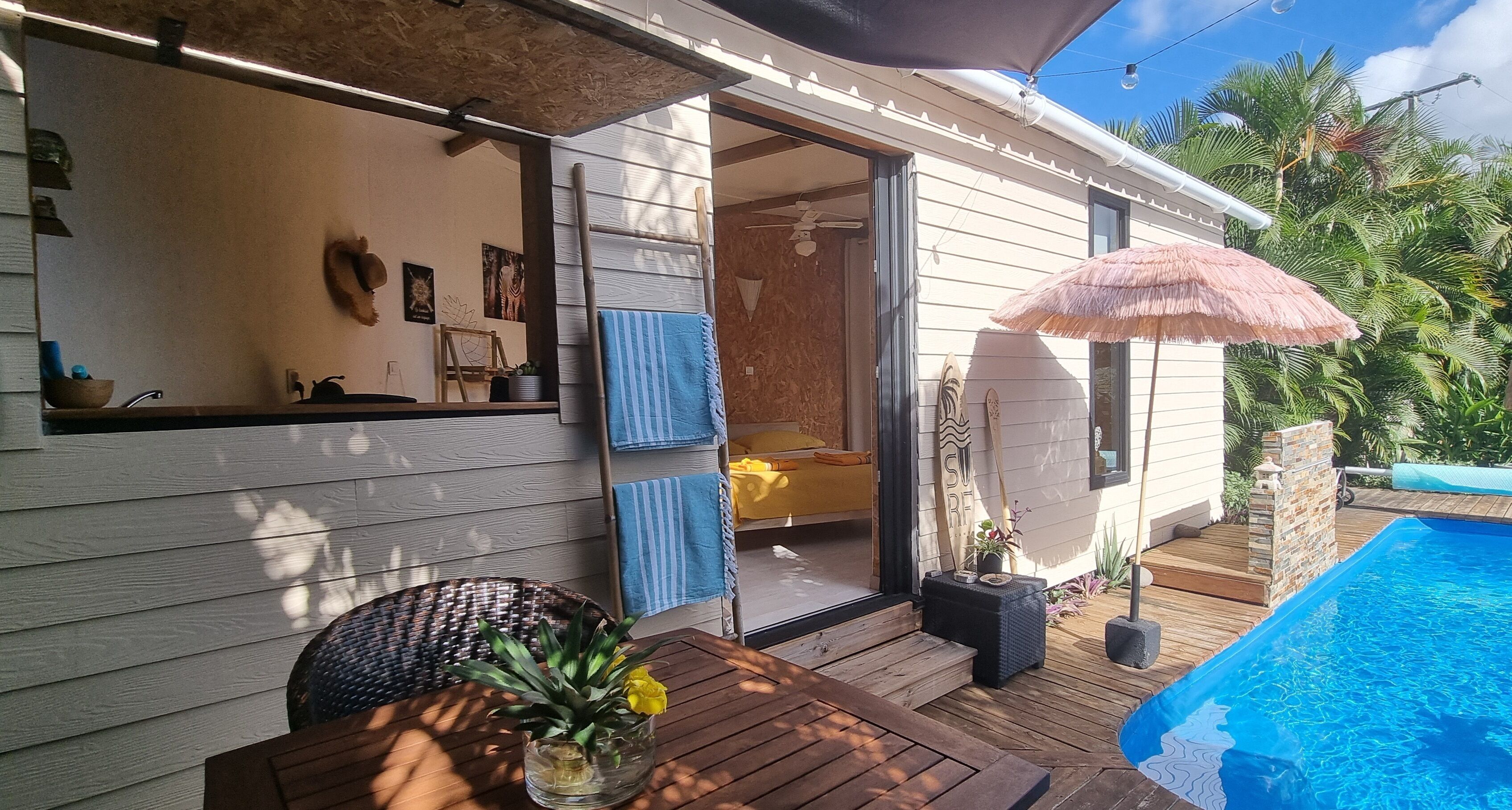 Une terrasse en bois avec une piscine, un parasol en chaume et une porte donnant sur une pièce avec une douche.