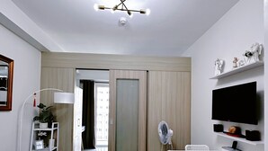 Una habitación moderna y bien iluminada con un sofá blanco, una mesa de comedor con sillas, un televisor montado en la pared y una lámpara en el techo.