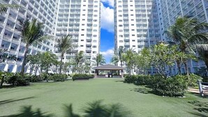 Un hotel moderno con dos edificios altos, un patio central con césped y palmeras, y un cielo azul despejado.