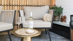 Un rincón al aire libre acogedor con un sofá acolchado, dos sillones, una mesita redonda y una botella de vino.