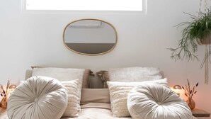 Una cama bien hecha con dos almohadones grandes y redondos, un espejo redondo en la pared y una planta colgante.