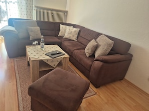 Un salon avec un canapé marron, un pouf assorti, une table basse recouverte d’une nappe à motifs et une bouteille d’eau.