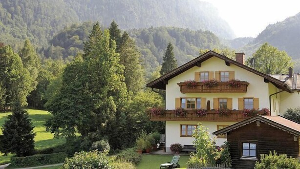 Un chalet a due piani con un balcone, immerso in un giardino curato e con montagne boscose sullo sfondo.