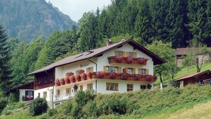 Un chalet a due piani con un balcone, immerso nel verde e con montagne sullo sfondo.