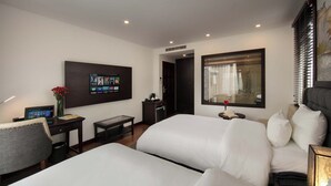 A hotel room with two beds, a desk with a computer, a small table with a vase of flowers, and a large window with blinds. - MIBEE Hotel (Hanoi)