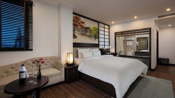 A hotel room with a bed, a sofa, a nightstand with a lamp, a mirror, and a painting on the wall. - MIBEE Hotel (Hanoi)