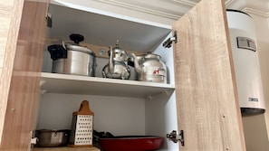 A kitchen cabinet with stainless steel pots, a red frying pan, and a wooden grater. - Cozy Apartment Near Rabat & Beach (Aïn Attig)