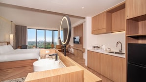 A modern hotel room with a large bed, a bathroom with a round mirror, a kitchen area, and a view of the city.