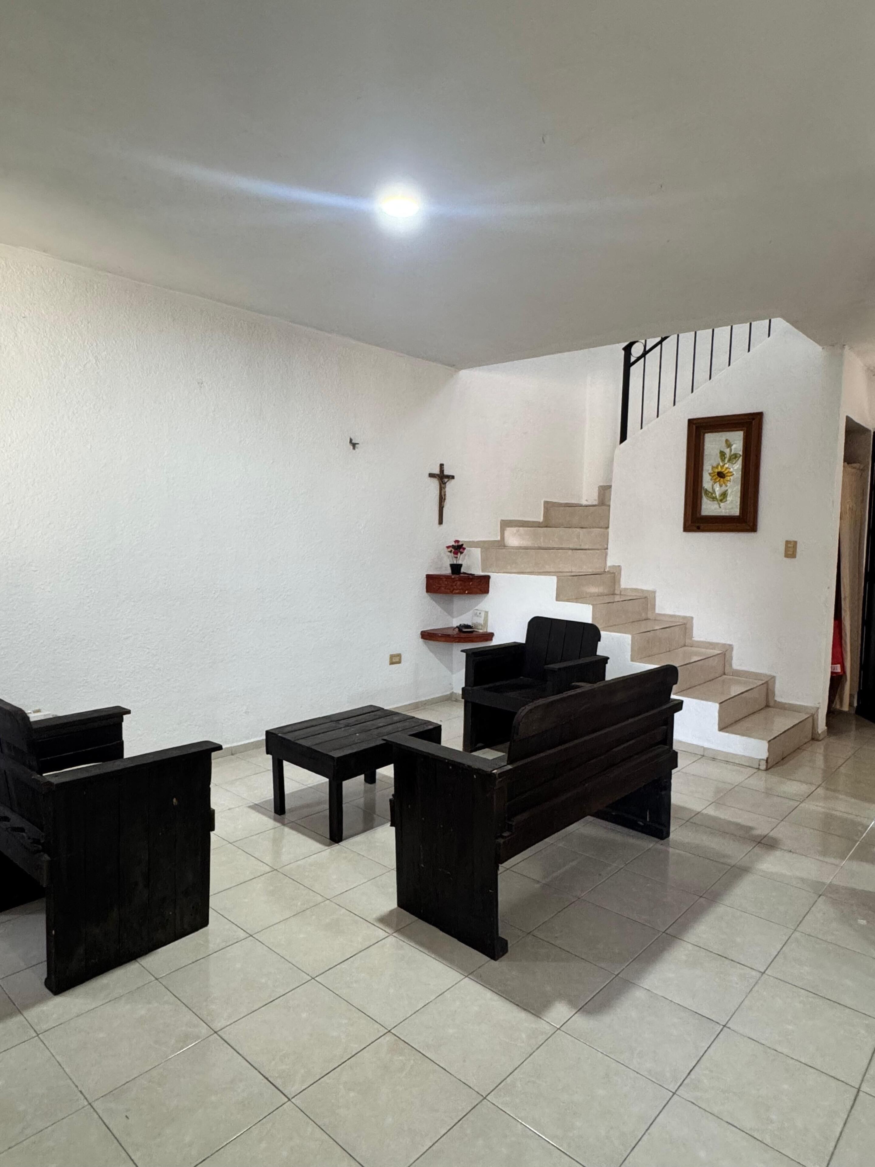 A spacious living area with a staircase leading to an upper floor, a wooden bench, a small table, and a wall-mounted cross.