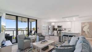 A modern living room with a large sofa, a coffee table, and a dining area with chairs. There is a kitchen with white cabinets and a large island. A balcony with a view of the water is visible.