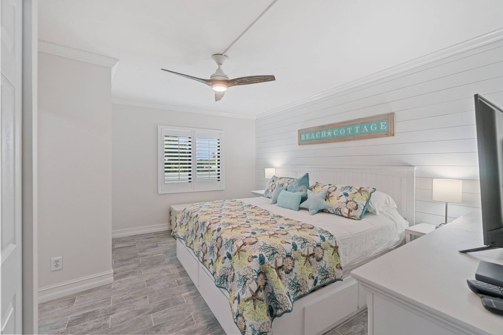 A bedroom with a bed, a ceiling fan, a window with blinds, a sign reading "BEACH COTTAGE", and a TV on a stand.