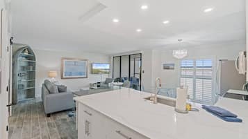 A modern kitchen with a marble countertop, a built-in sink, and a chandelier. There is a dining area with a glass table and chairs, and a living room with a gray sofa and a television.