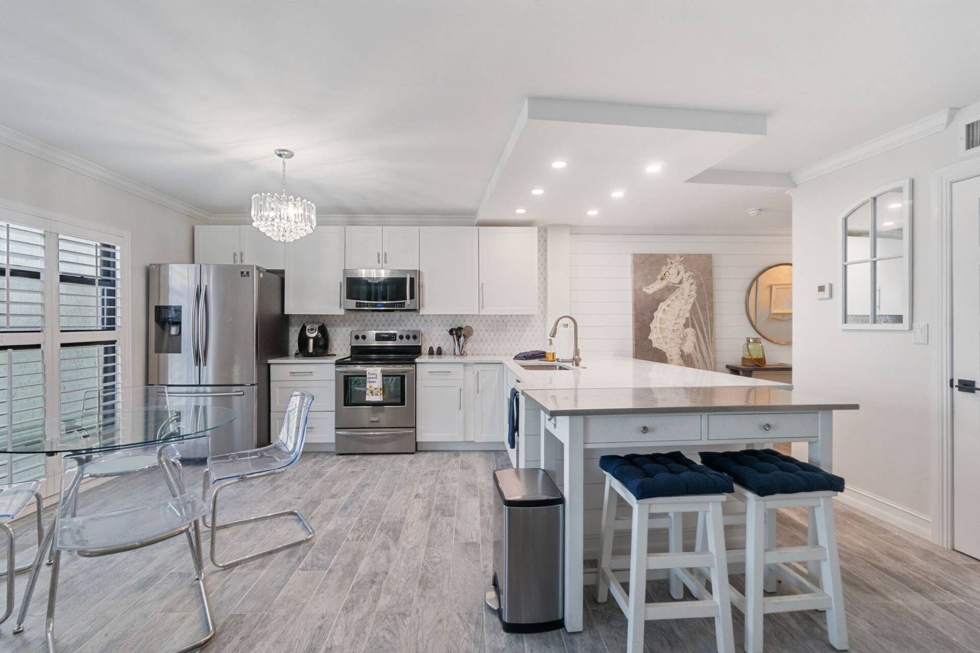 A modern kitchen with white cabinetry, stainless steel appliances, a glass dining table, and a chandelier.