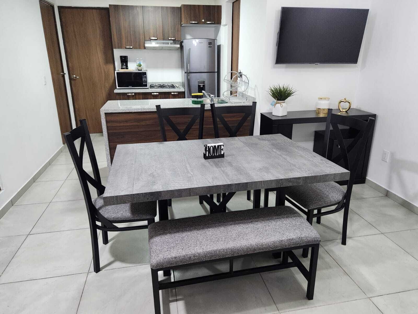 Un comedor con una mesa gris, sillas negras y un banco. Se ve una cocina al fondo con nevera, microondas y fogones.