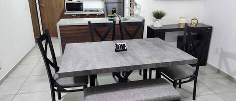 Un comedor con una mesa gris, sillas negras y un banco. Se ve una cocina al fondo con nevera, microondas y fogones.