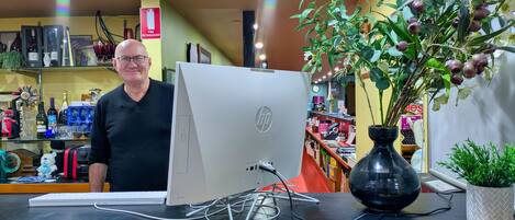 Un homme se tient derrière un bureau sur lequel se trouve un ordinateur de bureau, un vase avec une plante, et des étagères remplies de divers objets en arrière-plan.