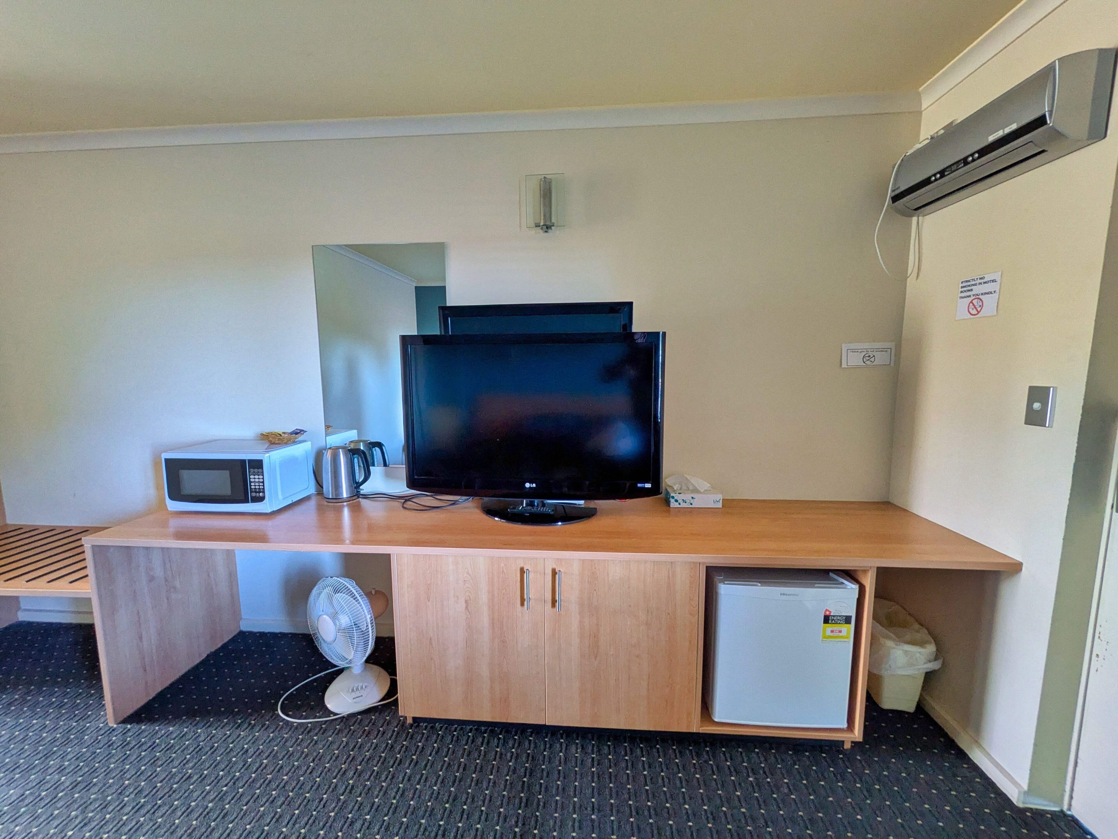 A compact hotel room with a wooden desk, a television, a microwave, a small refrigerator, and a fan.