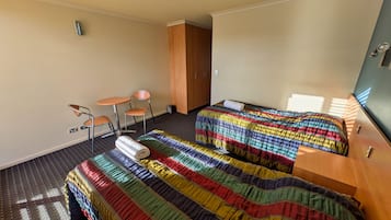A hotel room with two beds, a small table, two chairs, a wooden wardrobe, and a window with blinds.