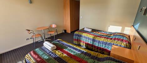 A hotel room with two beds, a small table, two chairs, a wooden wardrobe, and a window with blinds.
