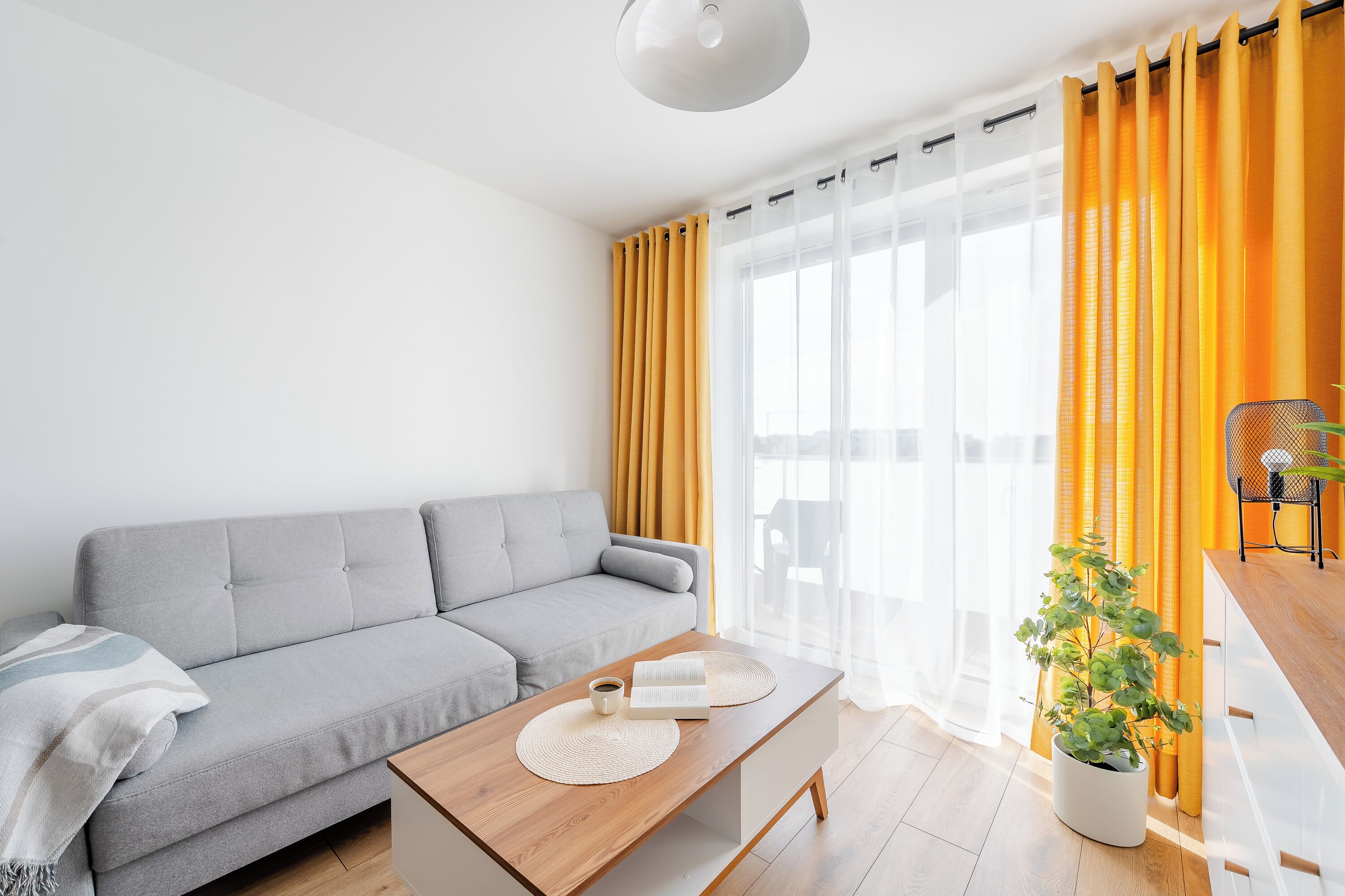 A modern living room with a grey sofa, a wooden coffee table, a potted plant, and a yellow curtain.