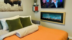 A hotel room with a bed, a mounted TV, a wall-mounted shelf with books, a framed picture, and a decorative plant. - Modern Downtown Suite - Walking Distance to Restaurants, Shops, & Grocery Stores (Golden)